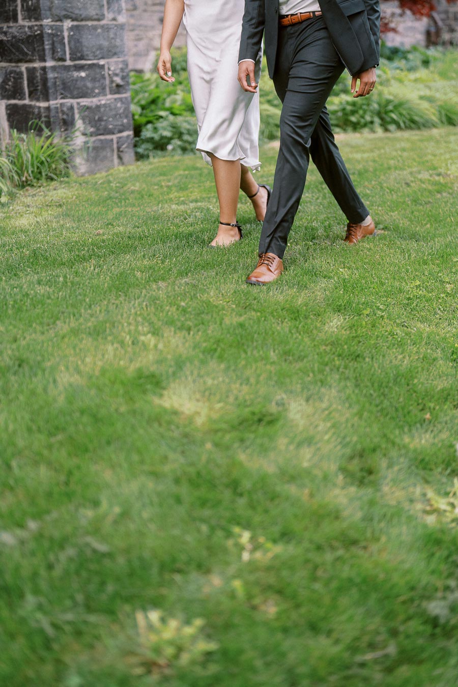 Two people dressed formally are walking side by side on a green lawn near a stone building at the University of Toronto.