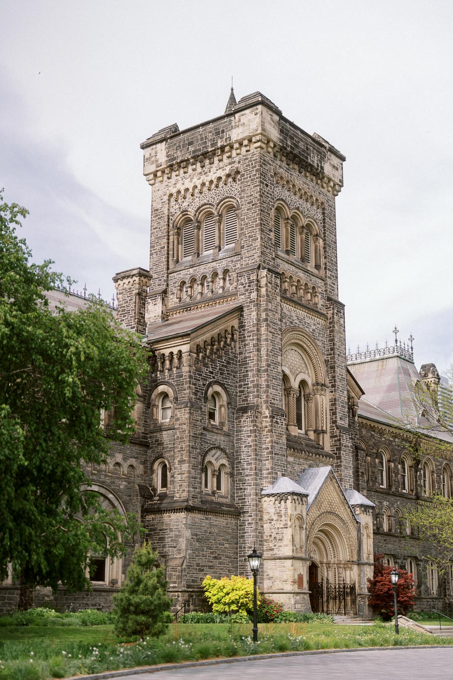 A historic stone tower with arched windows and an ornate entrance at the University of Toronto St. George campus, with trees and landscaped greenery in the foreground.