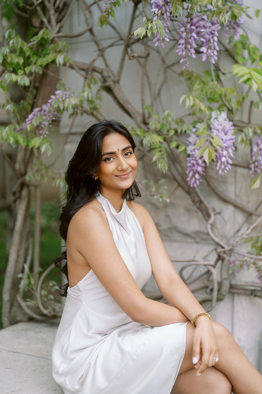 A woman in a white dress sits on a stone bench, framed by blooming purple wisteria vines at the University of Toronto.