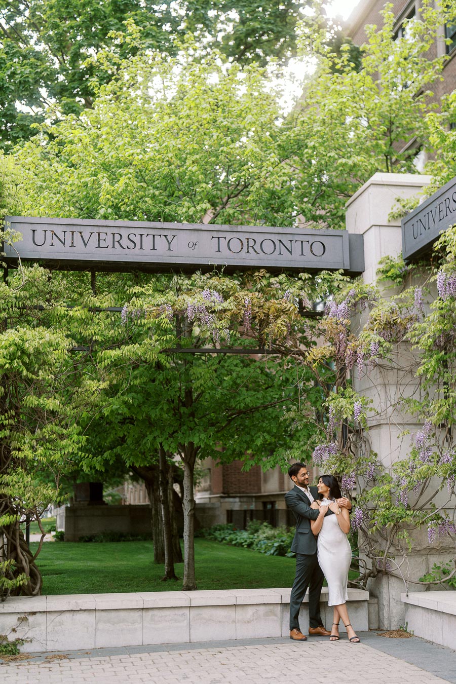 A couple poses together under a University of Toronto archway, with lush green trees and purple wisteria flowers in the background.