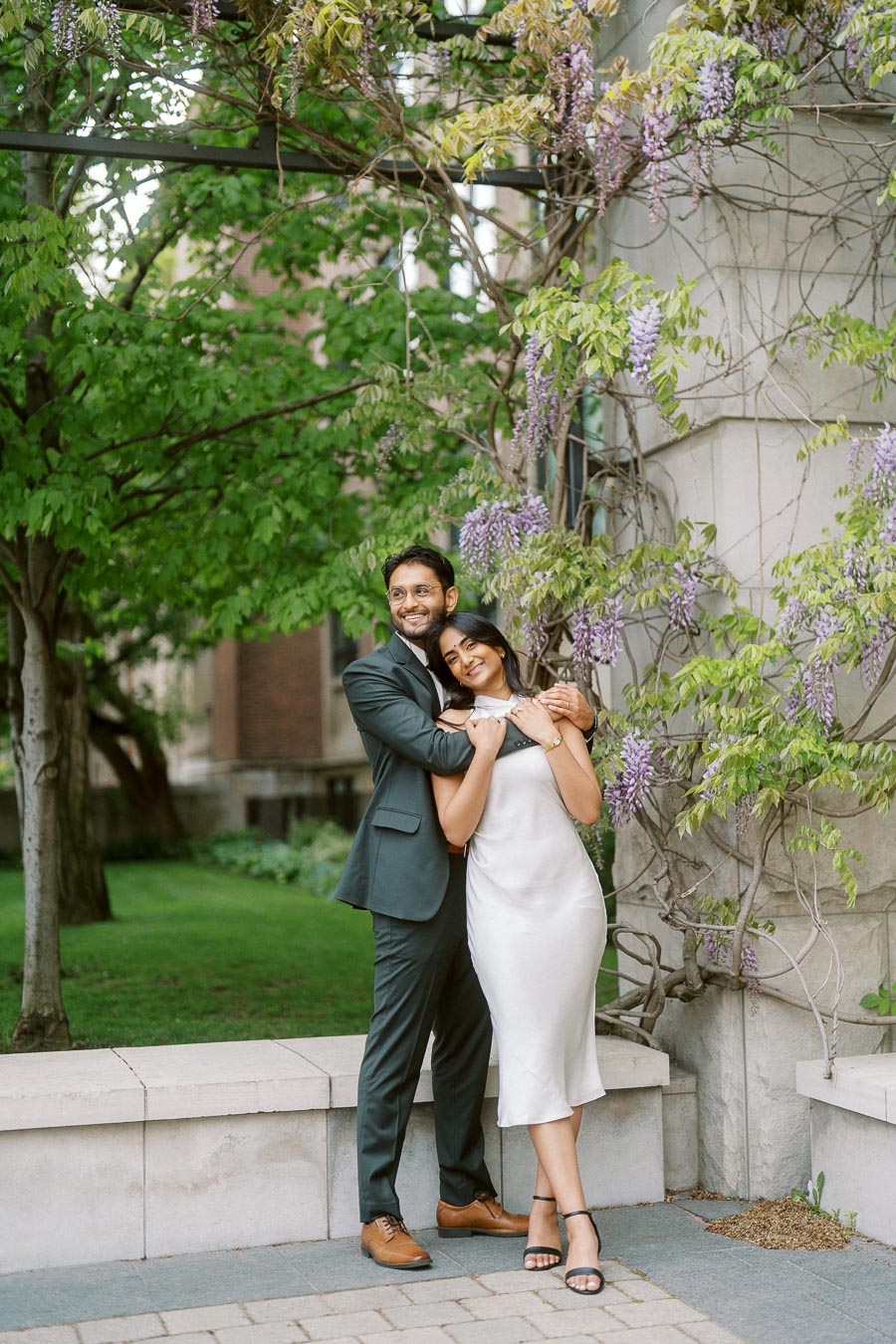 Tej and Priyana stand together, smiling and embracing, next to a stone wall covered in purple spring blossoms at the University of Toronto.