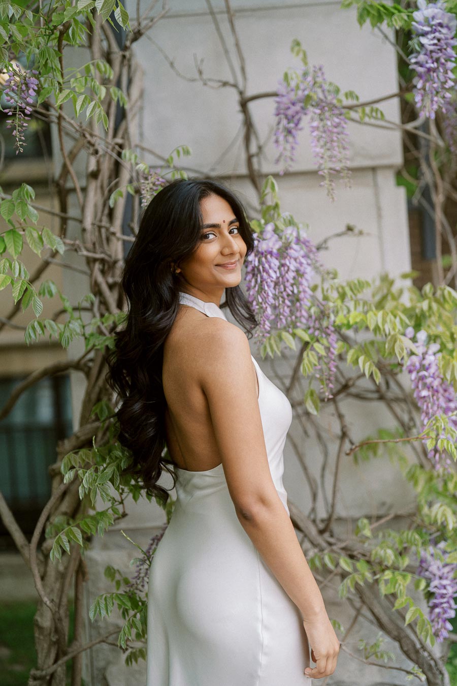 A woman in a white dress stands beside wisteria blossoms and greenery at the University of Toronto St. George campus.