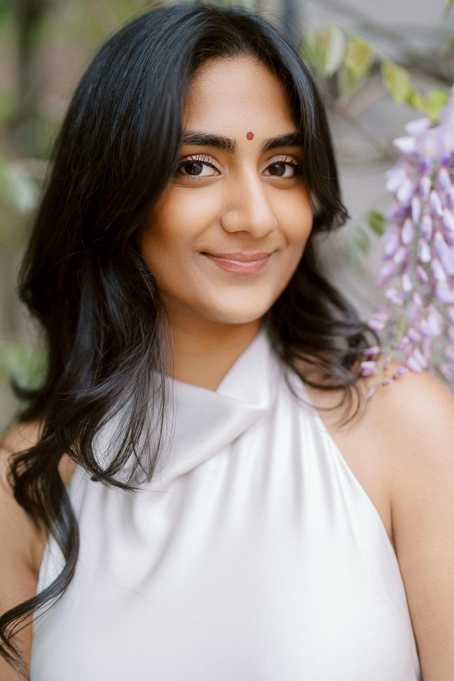 Priyana smiles in a close-up portrait with dark hair and a white dress, standing near purple spring blossoms at the University of Toronto.
