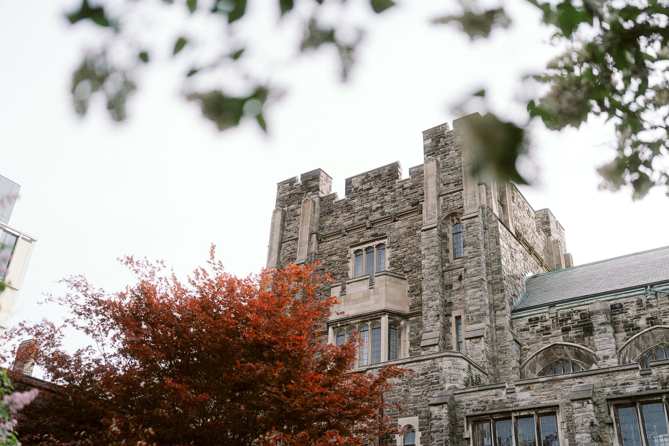 A historic stone building with Gothic windows at the University of Toronto St. George Campus, framed by leafy branches and red foliage.