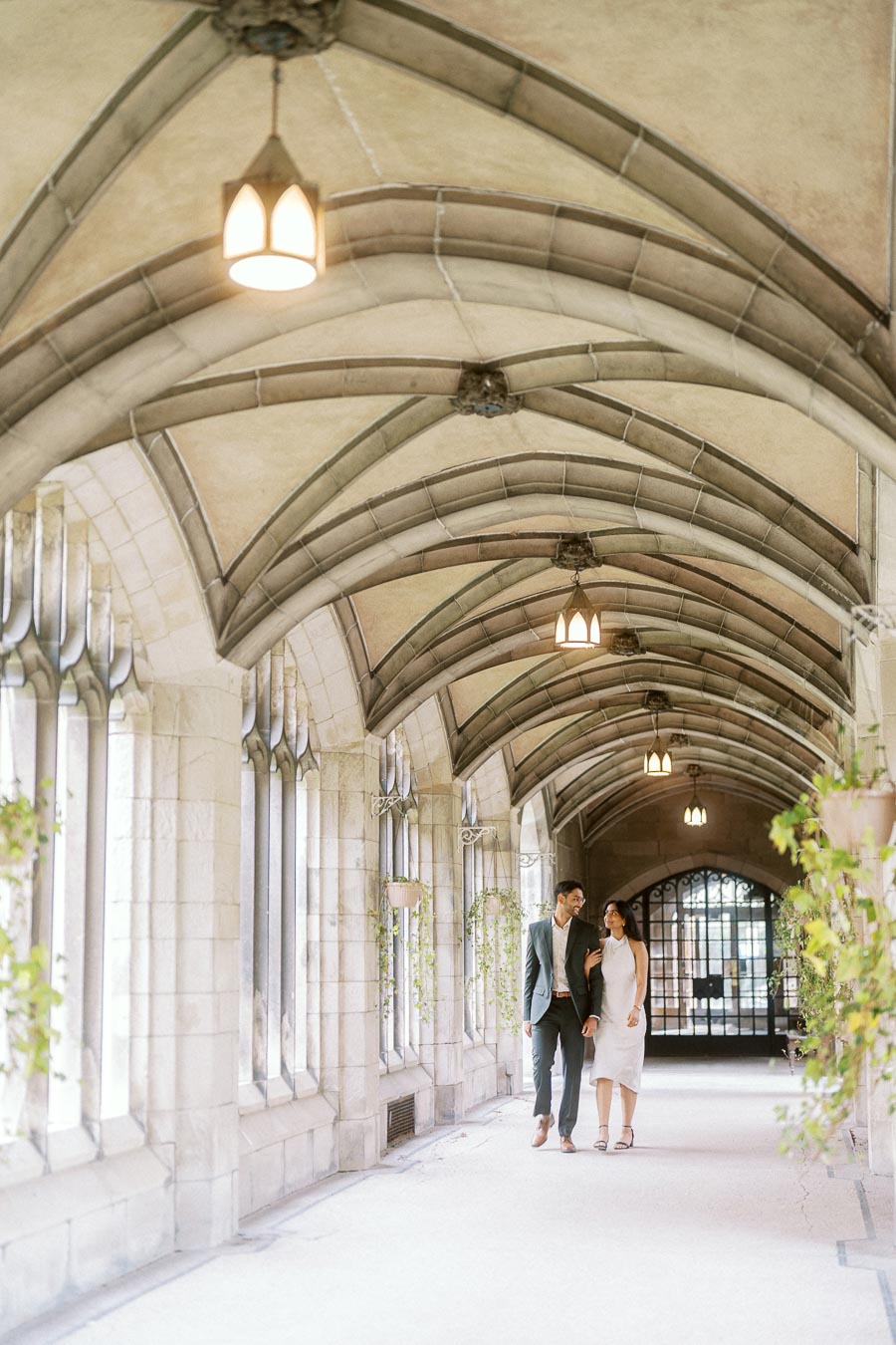 Tej and Priyana walk together under a vaulted stone archway at the University of Toronto, surrounded by greenery and historic architecture.