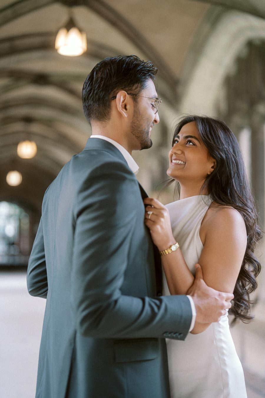 Tej and Priyana stand close together under a stone archway at the University of Toronto, looking at each other and smiling.