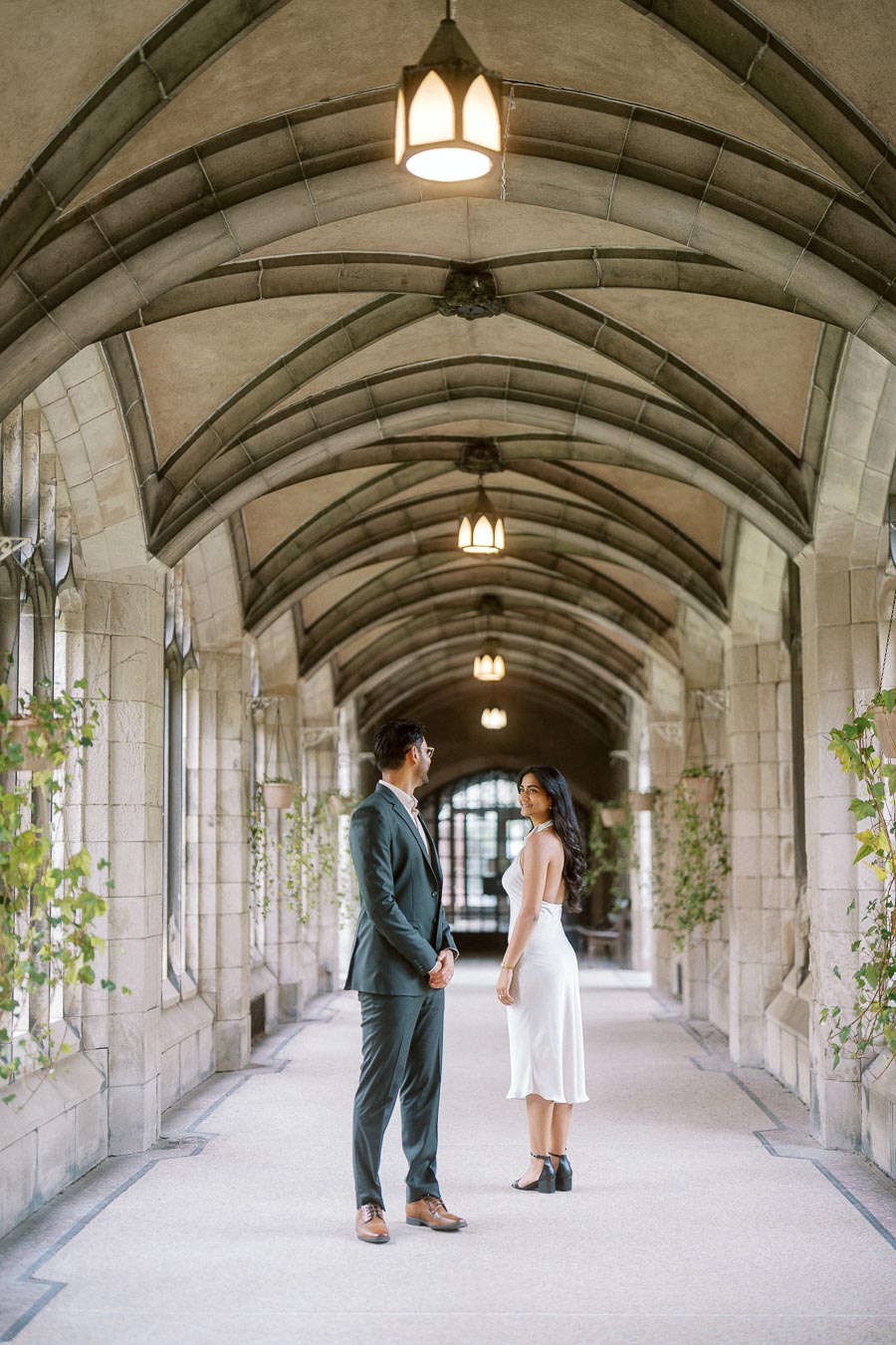 A couple stands facing each other in a stone Gothic archway corridor at the University of Toronto, with greenery along the walls.