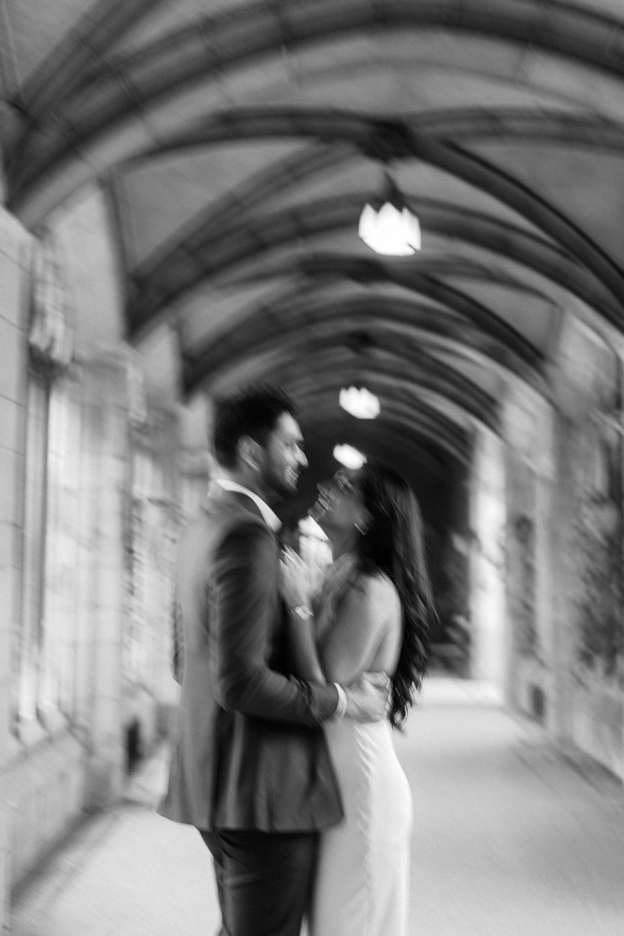 A blurred black and white photo of a couple standing together under a historic stone archway at the University of Toronto.