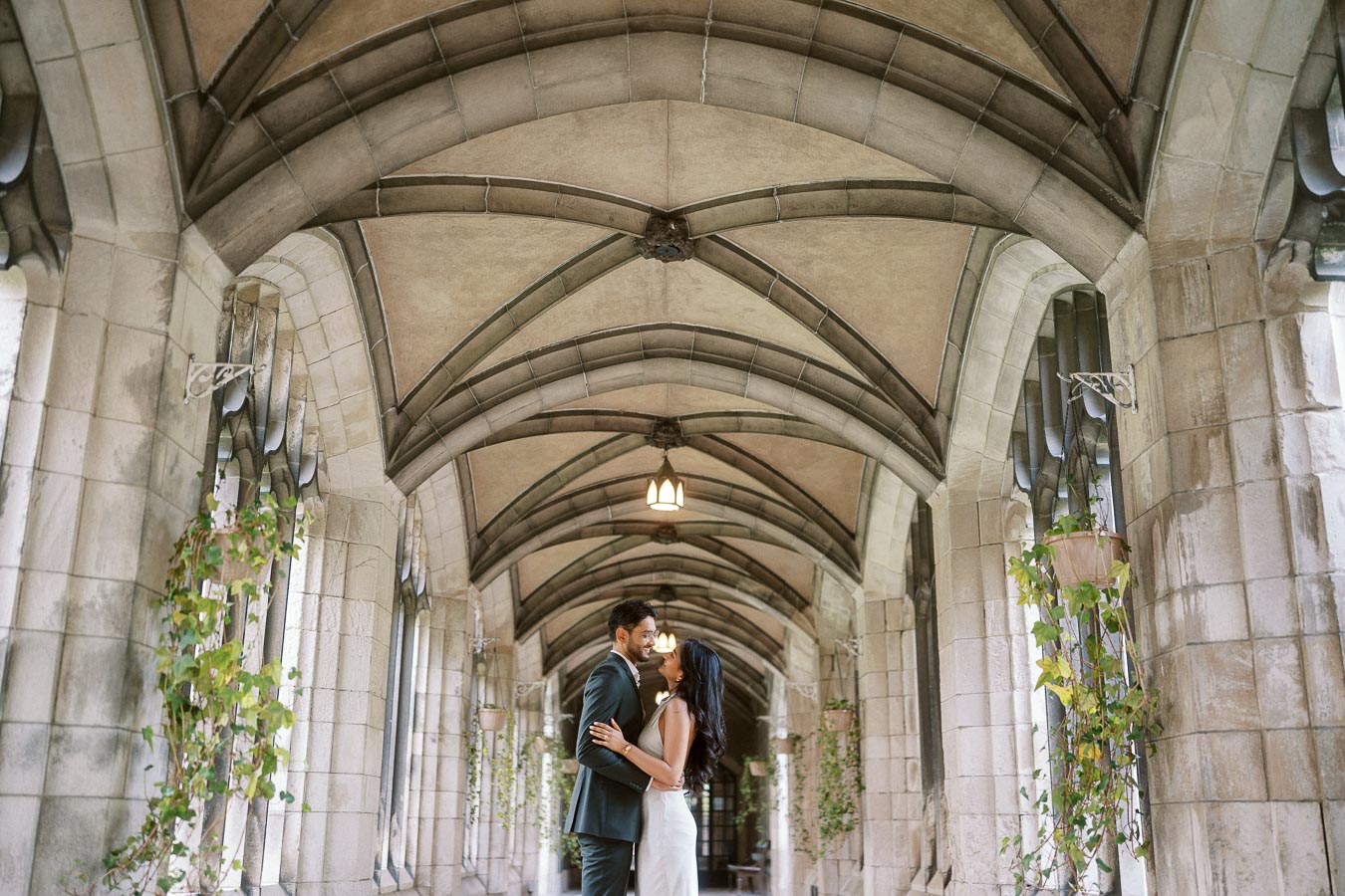 A couple stands embracing under a vaulted stone archway with hanging plants at the University of Toronto campus.