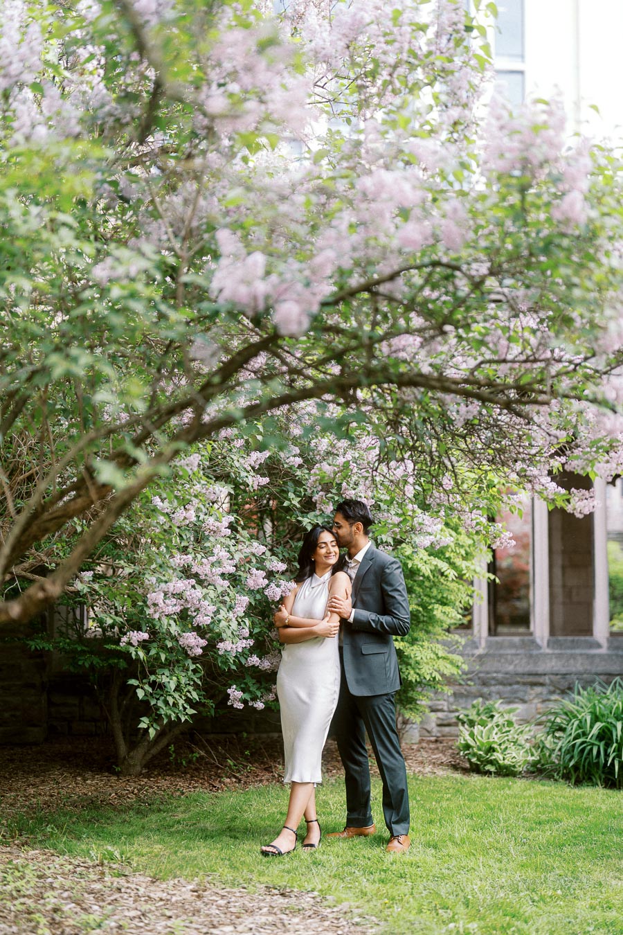 A couple stands close together under blooming lilac trees on the University of Toronto campus, surrounded by greenery and historic architecture.