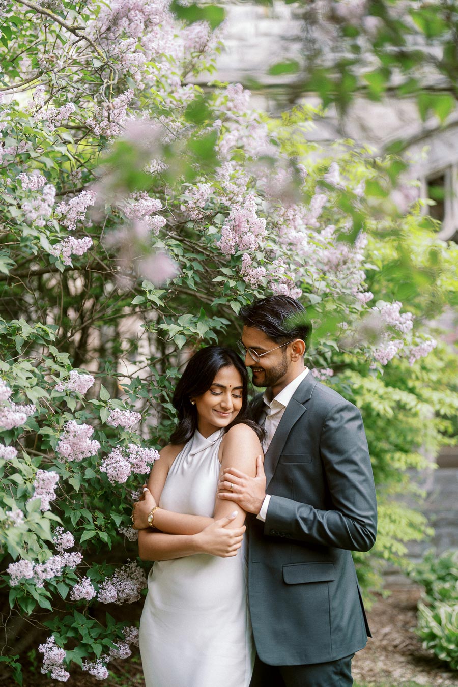 Tej and Priyana embrace beside blooming lilac bushes on the University of Toronto campus, with soft greenery and flowers surrounding them.