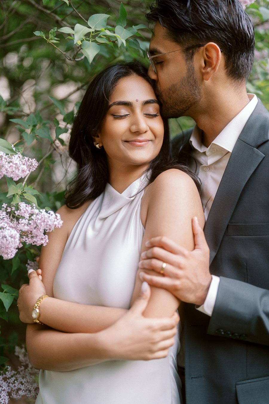 Tej gently kisses Priyana on the forehead while embracing her beside blooming lilac flowers at the University of Toronto campus.