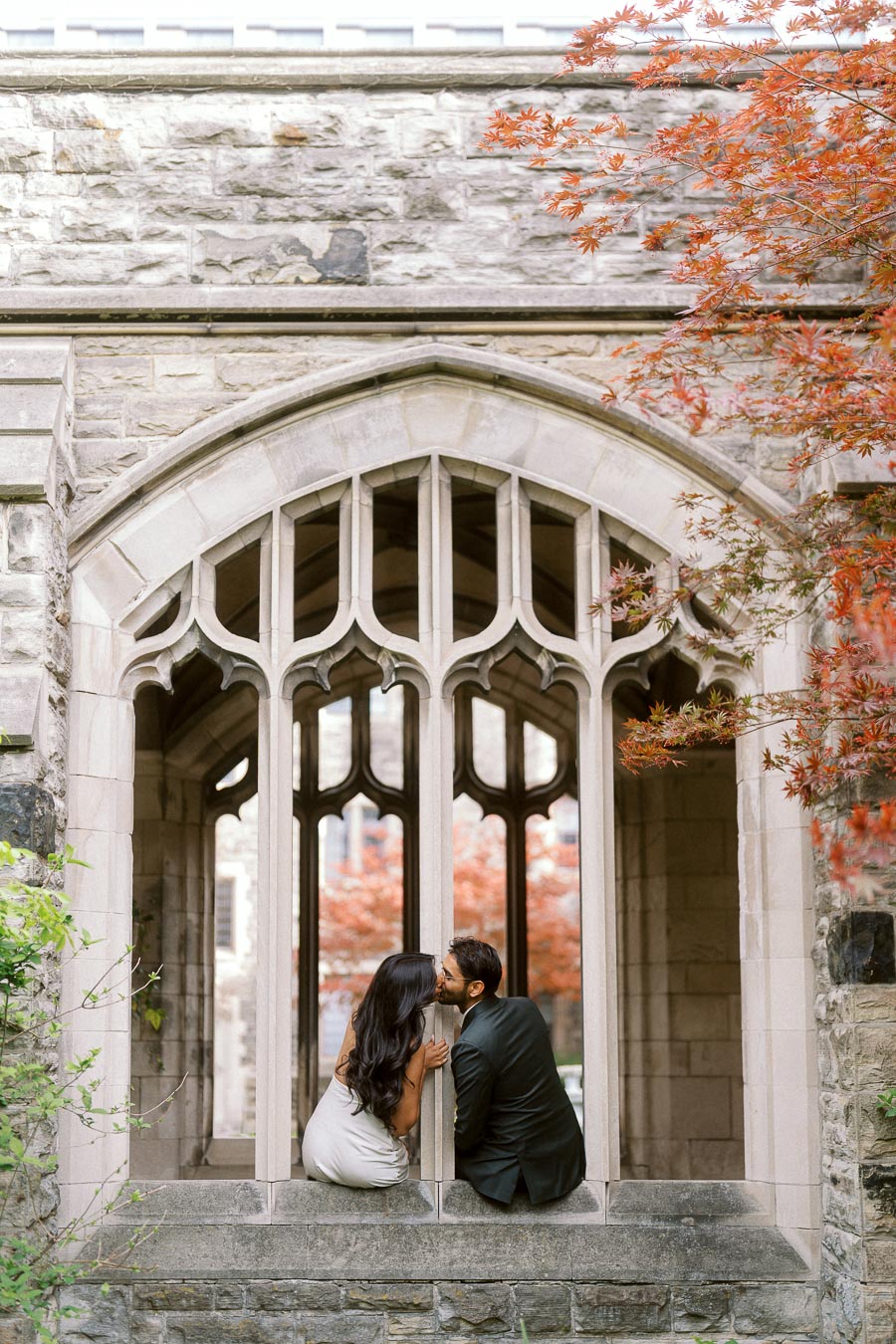 Tej and Priyana kiss while sitting in a stone Gothic archway at the University of Toronto, surrounded by leafy branches and historic architecture.