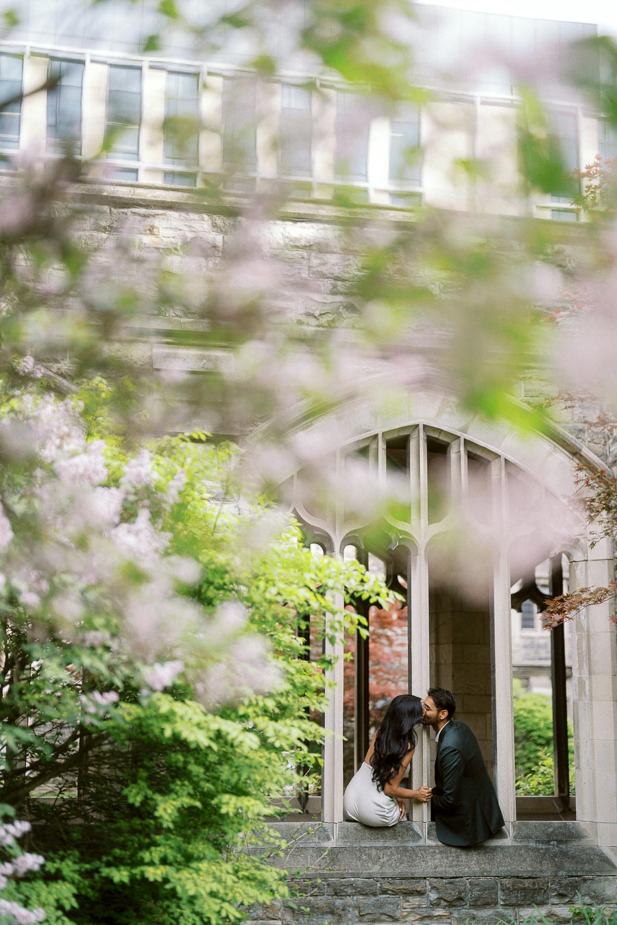 A couple sits in a stone archway surrounded by spring greenery and pink blossoms, sharing a kiss at the University of Toronto campus.