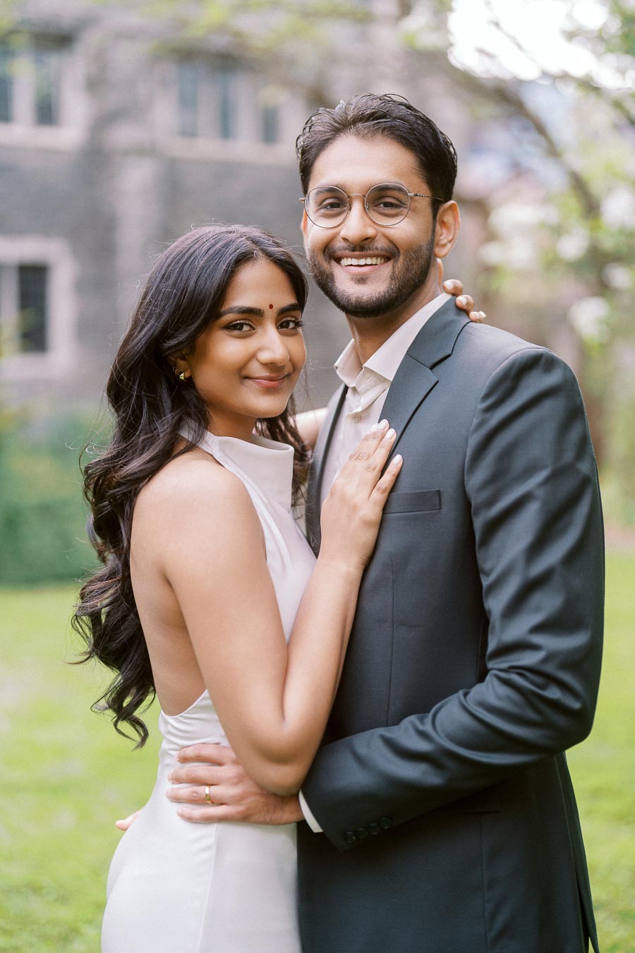 A couple stands closely together outdoors, smiling at the camera with historic stone building and greenery behind them at University of Toronto.