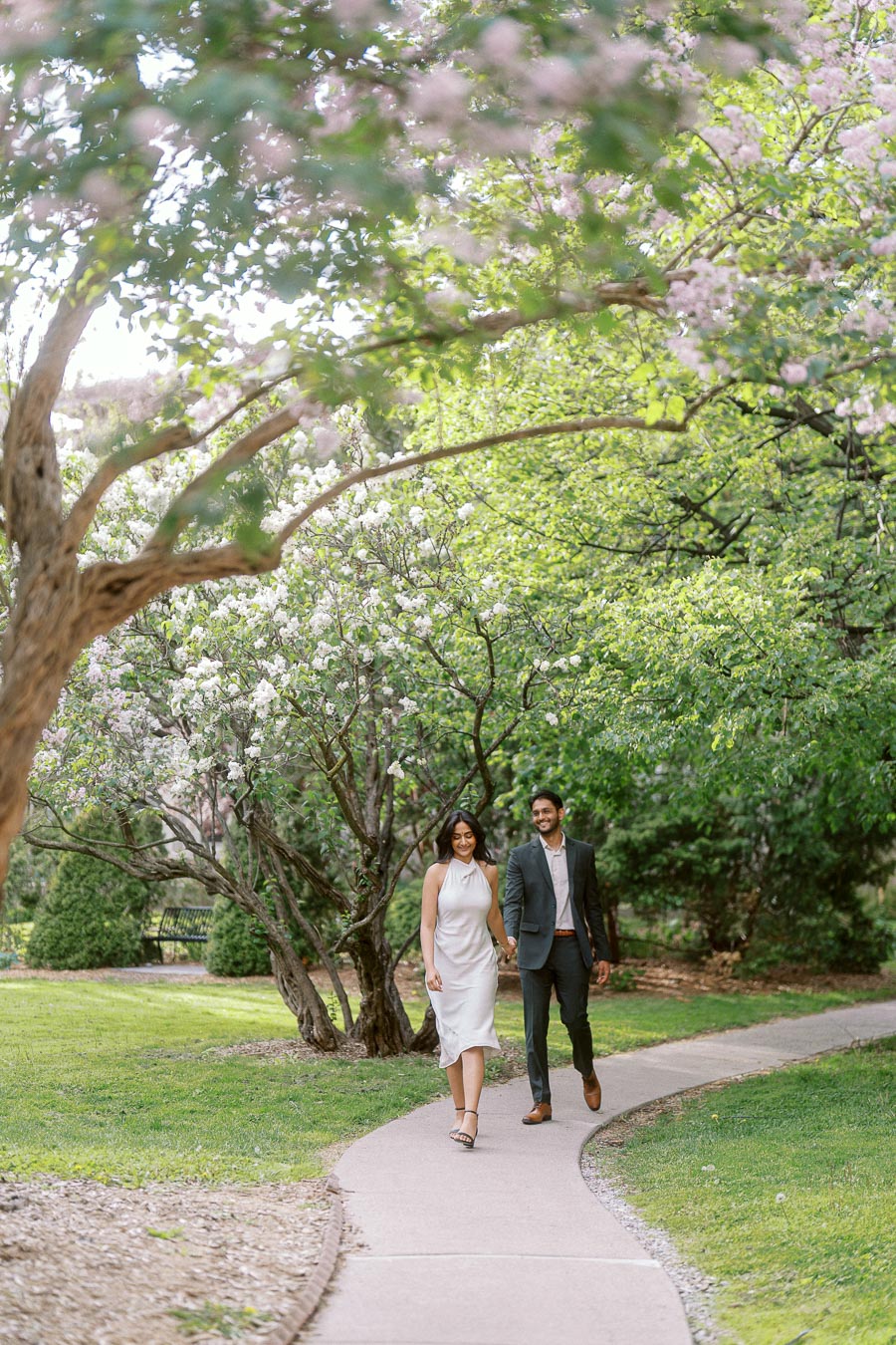 A couple walks hand in hand along a curved path lined with blossoming trees and greenery at the University of Toronto campus.