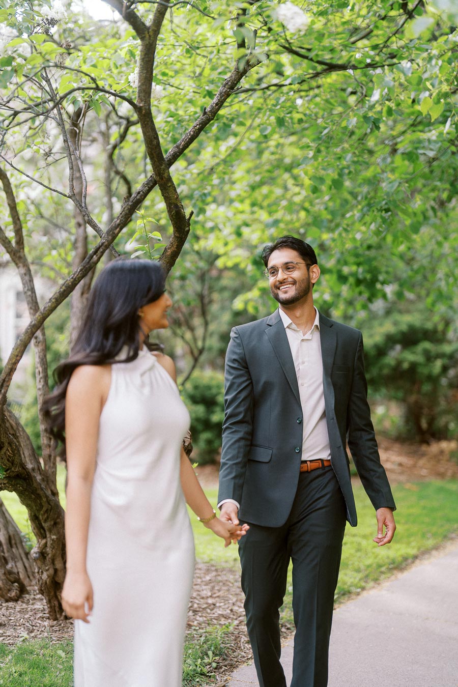 A couple walks hand in hand along a shaded campus path surrounded by green trees at the University of Toronto.