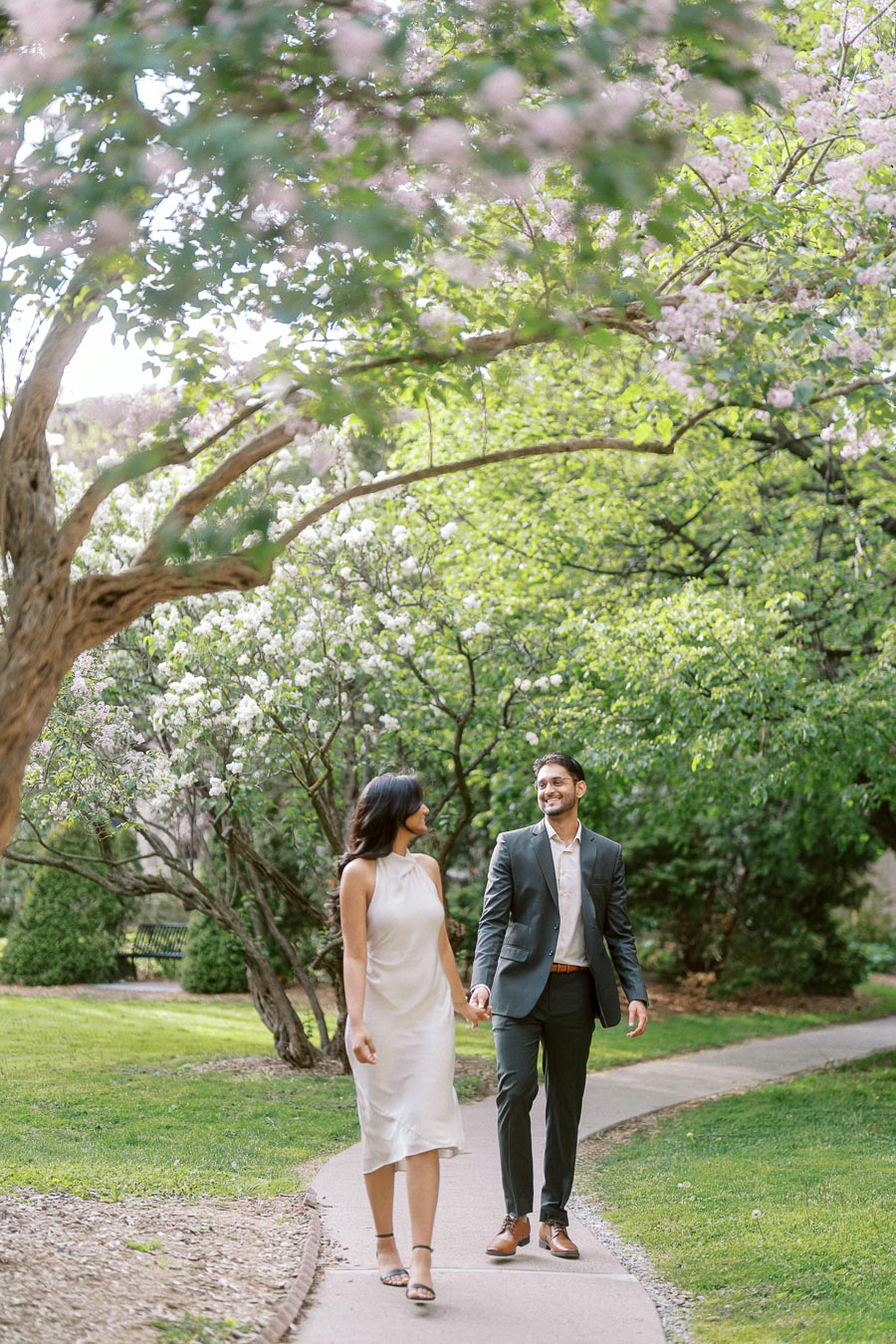 A couple walks hand in hand on a garden path under blooming trees at the University of Toronto campus.