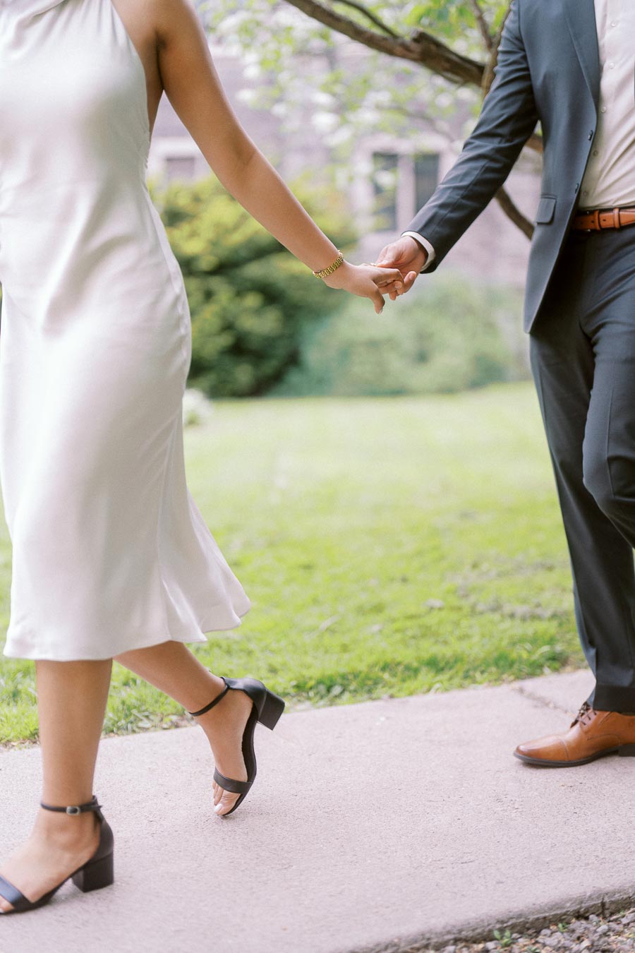 A couple holding hands and walking on a sidewalk at University of Toronto, with greenery and campus architecture in the background.