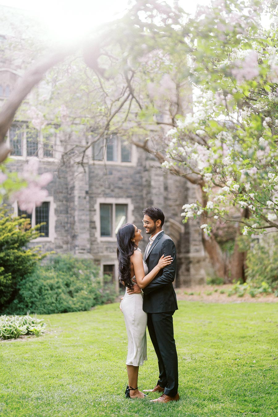 A couple stands close together on a green lawn under flowering trees with historic stone buildings in the background at the University of Toronto.