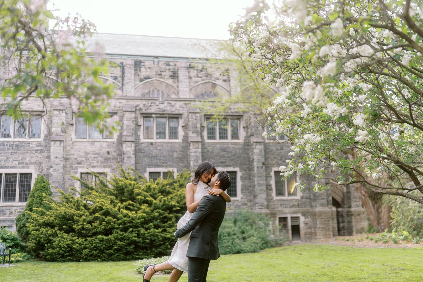 Tej lifts Priyana in a garden at the University of Toronto, surrounded by spring blossoms and historic stone buildings.