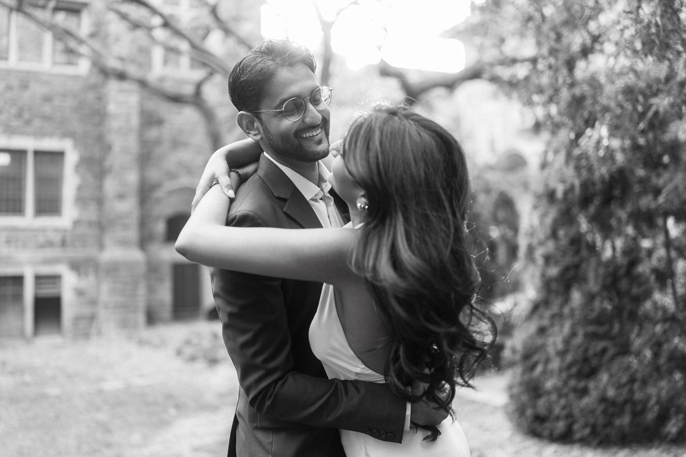 A couple stands close together, embracing and smiling, outdoors near a historic stone building at the University of Toronto campus.
