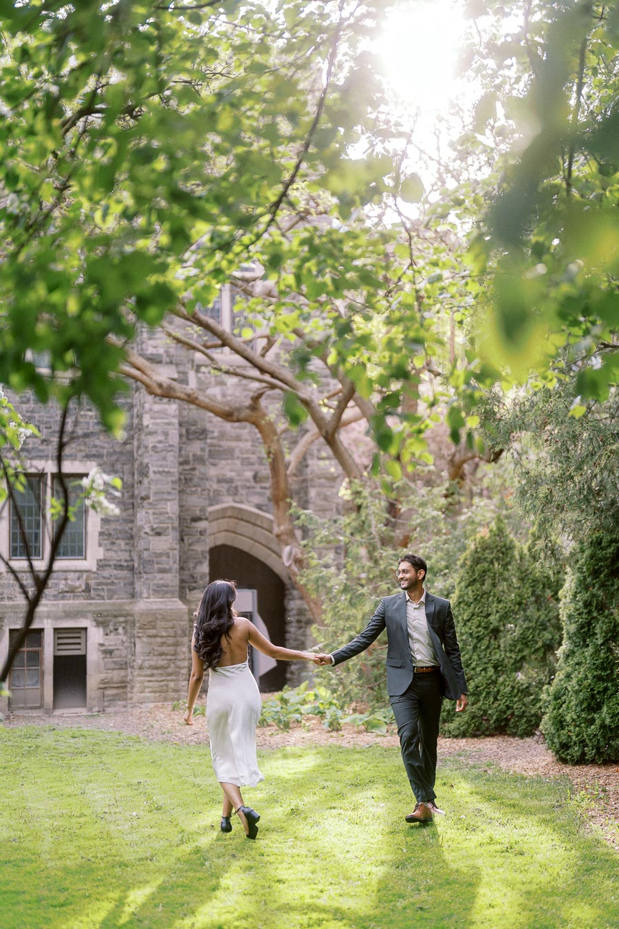 A couple walks hand in hand on a green lawn under leafy trees with stone university buildings in the background.