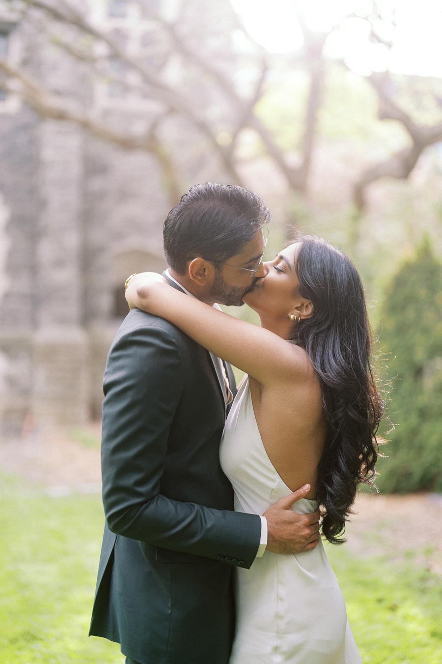 Tej and Priyana embrace and kiss outdoors on the University of Toronto campus with stone buildings and greenery in the background.