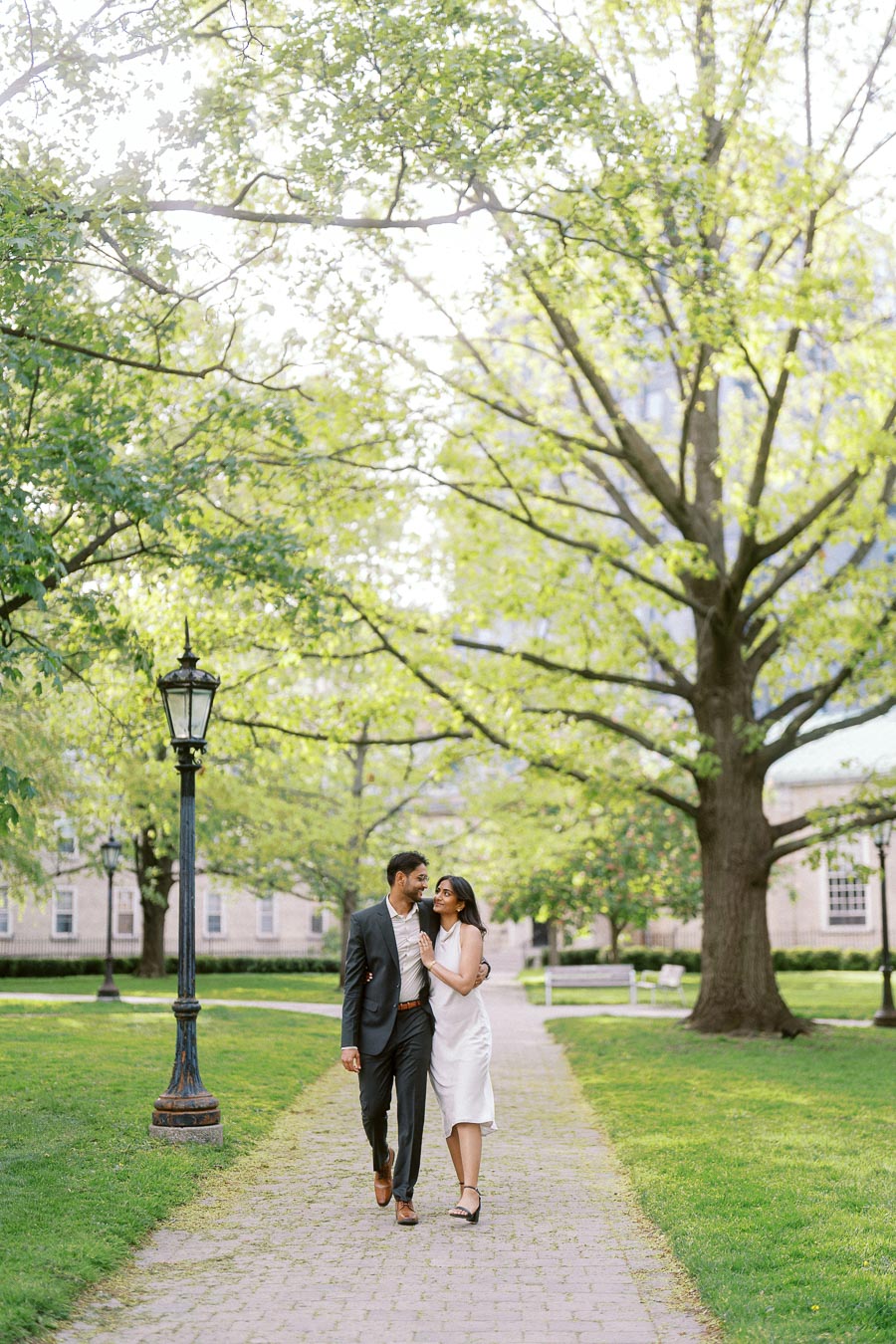 A couple walks together on a tree-lined path at the University of Toronto St. George campus, with green grass and historic buildings in the background.