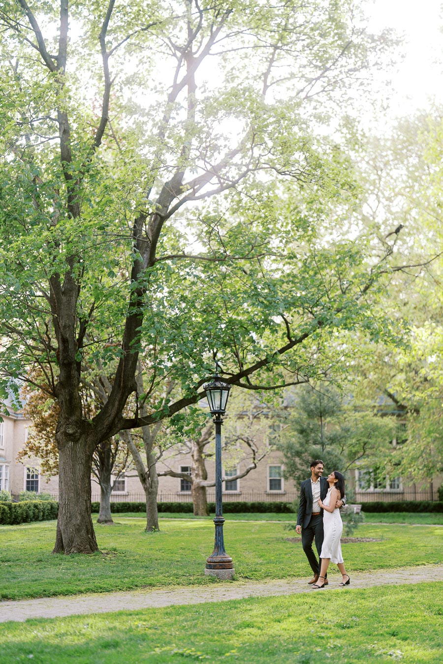 A couple walks arm in arm along a path under large green trees on the University of Toronto campus.