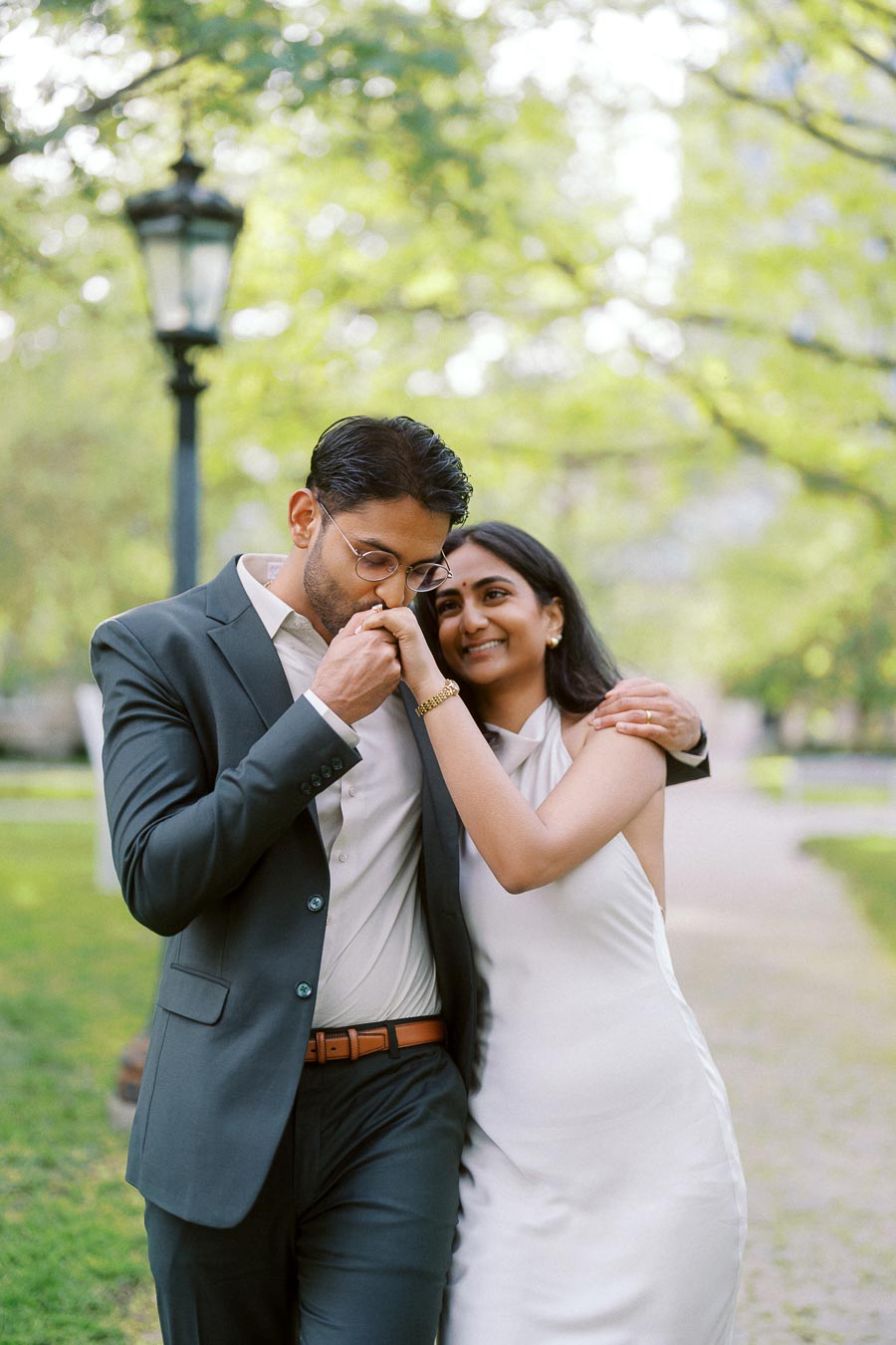 Tej and Priyana walk together outdoors at the University of Toronto, with Tej kissing Priyana's hand and greenery in the background.