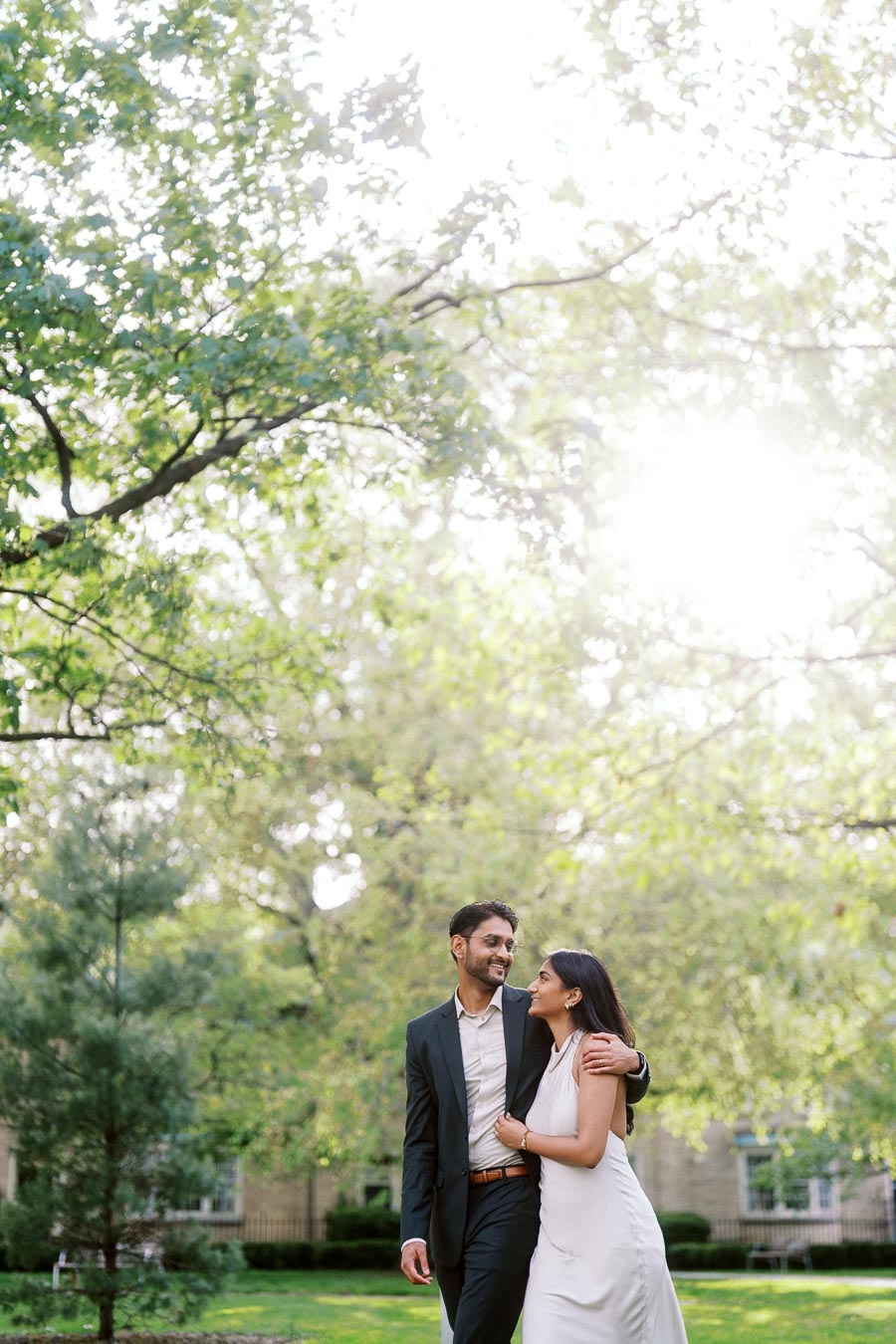 A couple, Tej and Priyana, walk together on a green campus lawn surrounded by leafy trees at the University of Toronto.