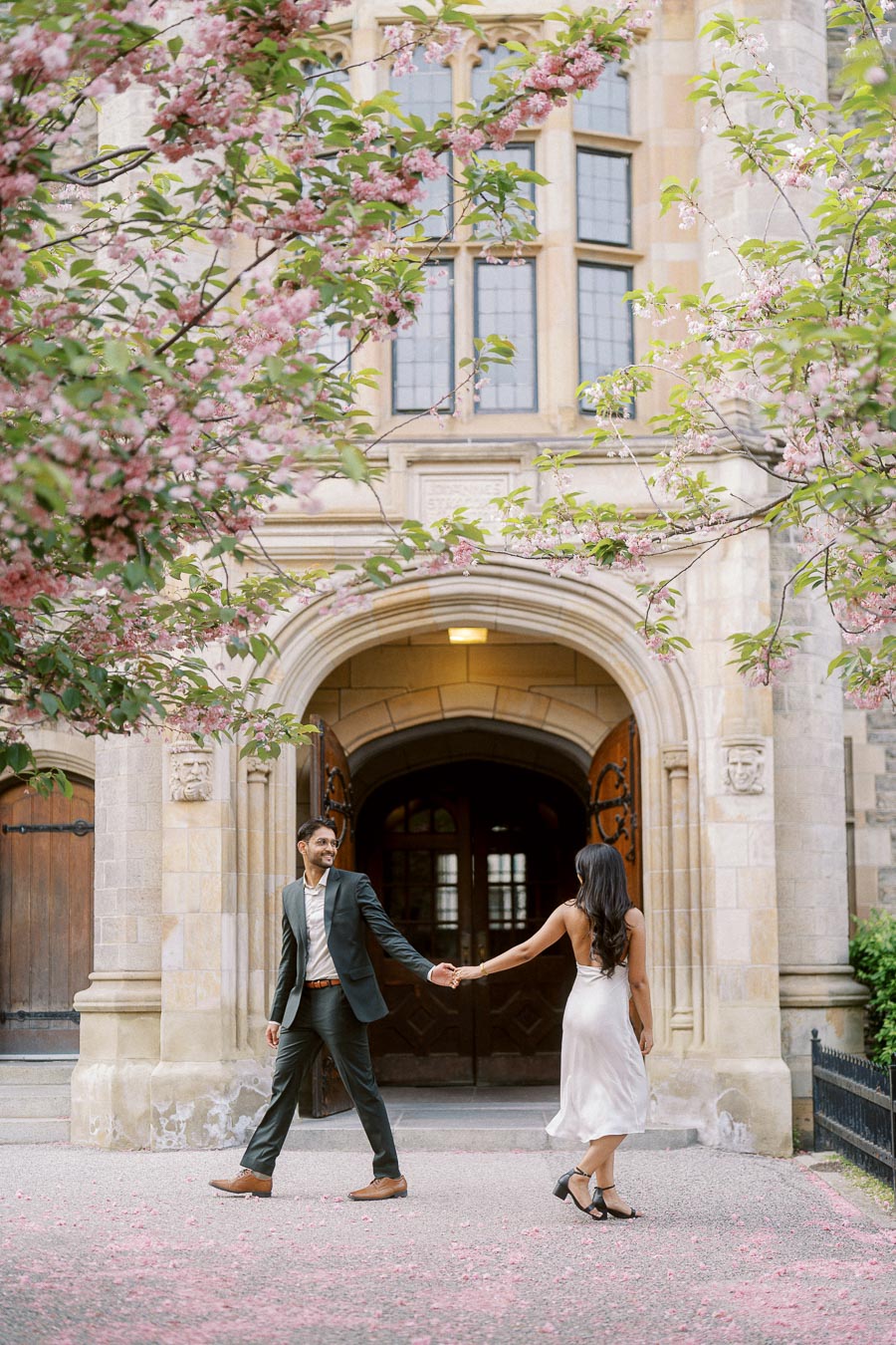 A couple holding hands and walking under pink spring blossoms in front of a historic stone archway at the University of Toronto.