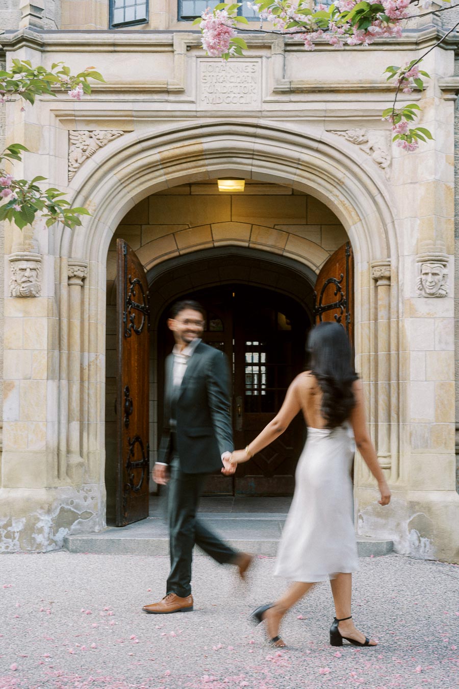 A couple walks hand in hand past a large stone archway with wooden doors at the University of Toronto, surrounded by pink spring blossoms.