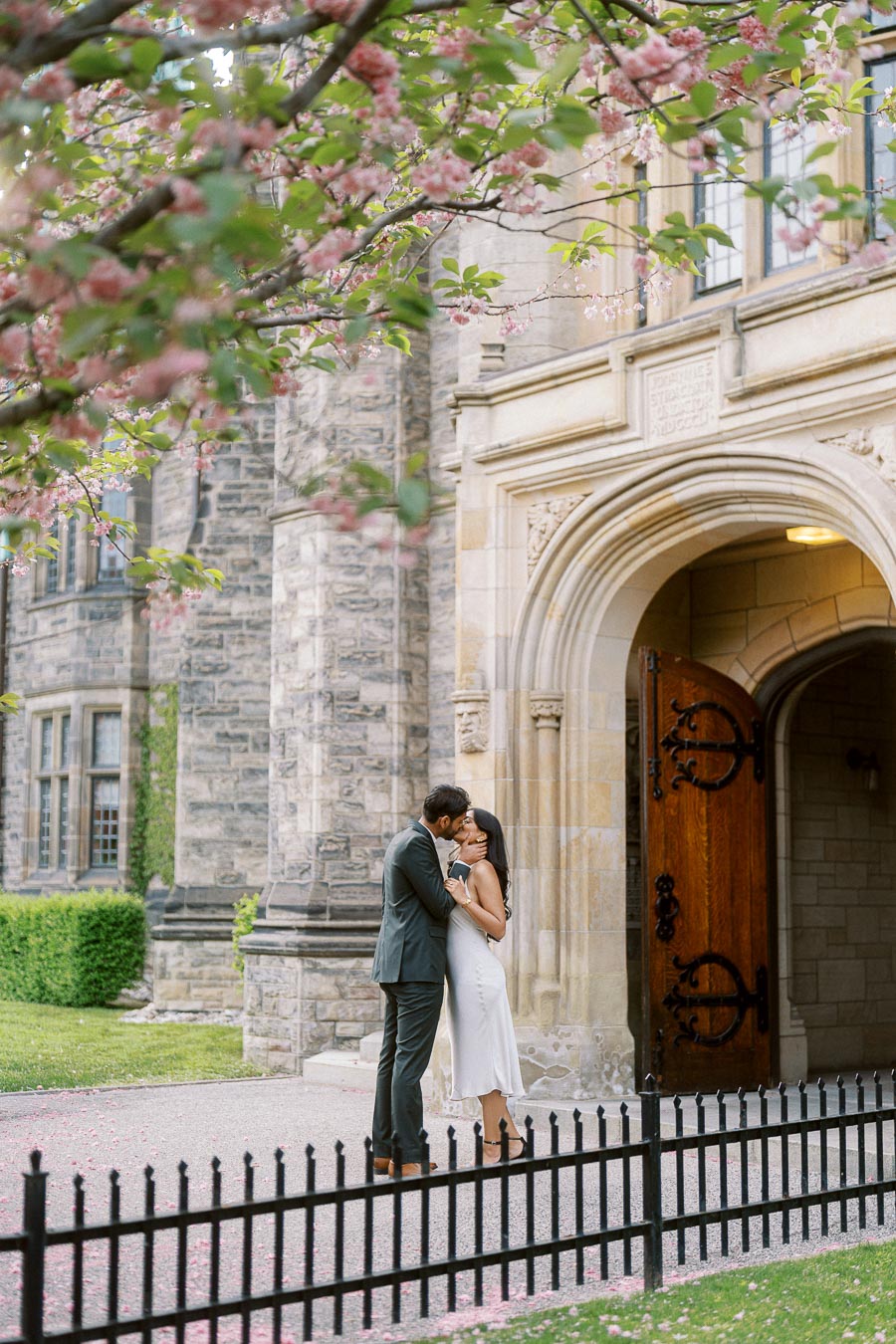 A couple stands embracing and kissing beneath pink spring blossoms in front of a historic stone archway at the University of Toronto.