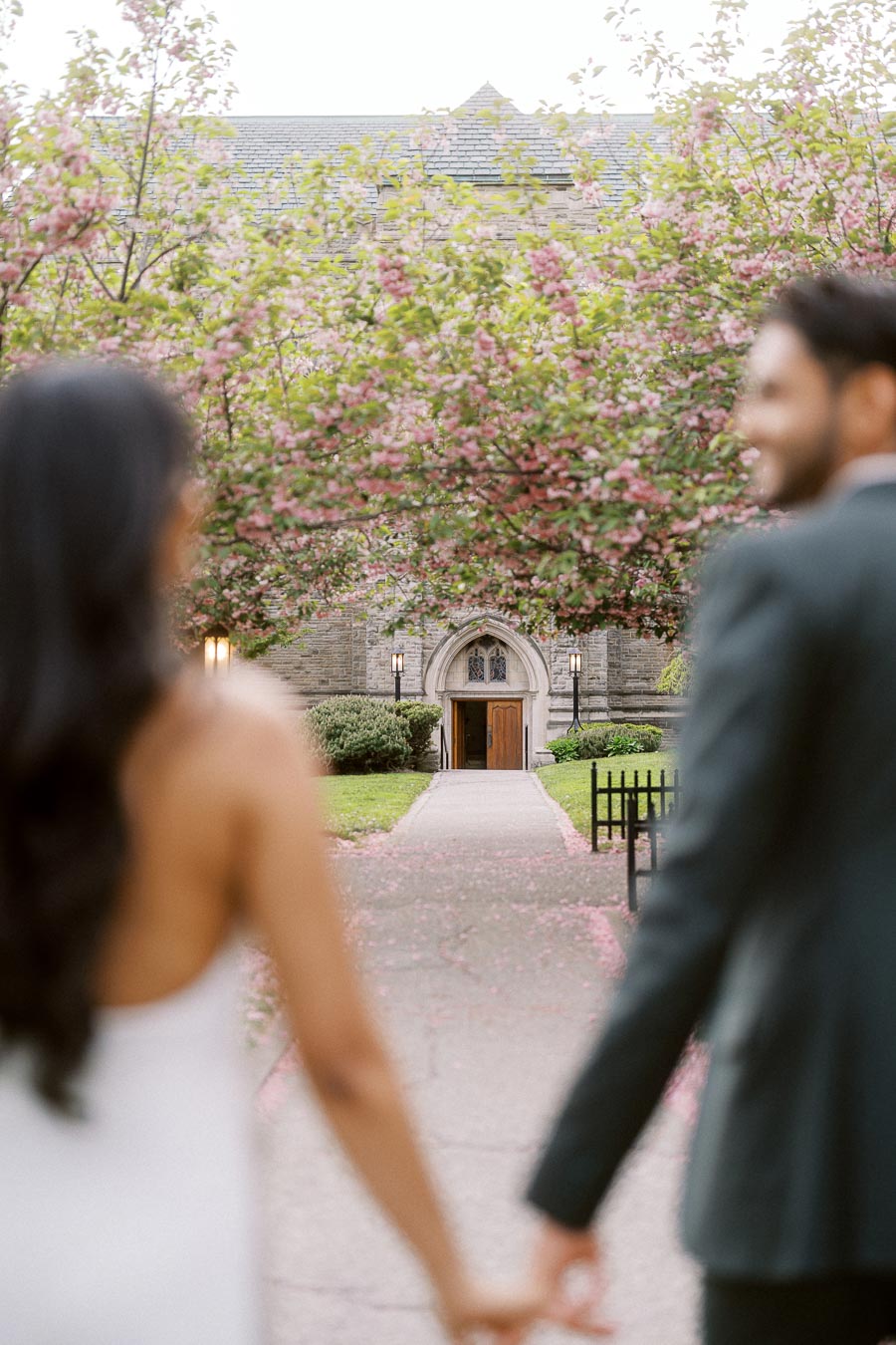 A couple holding hands in the foreground, walking on a flower-lined path toward a stone archway at the University of Toronto.