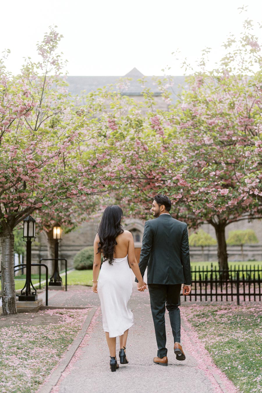 A couple walks hand in hand down a path lined with pink spring blossoms at the University of Toronto campus.