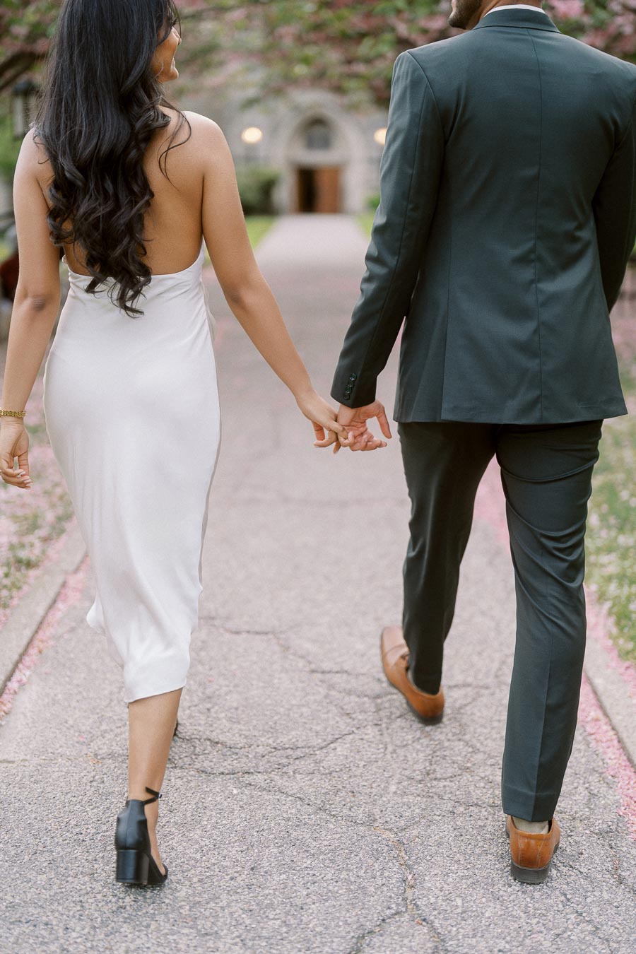 A couple walks hand in hand down a tree-lined path at the University of Toronto, photographed from behind.