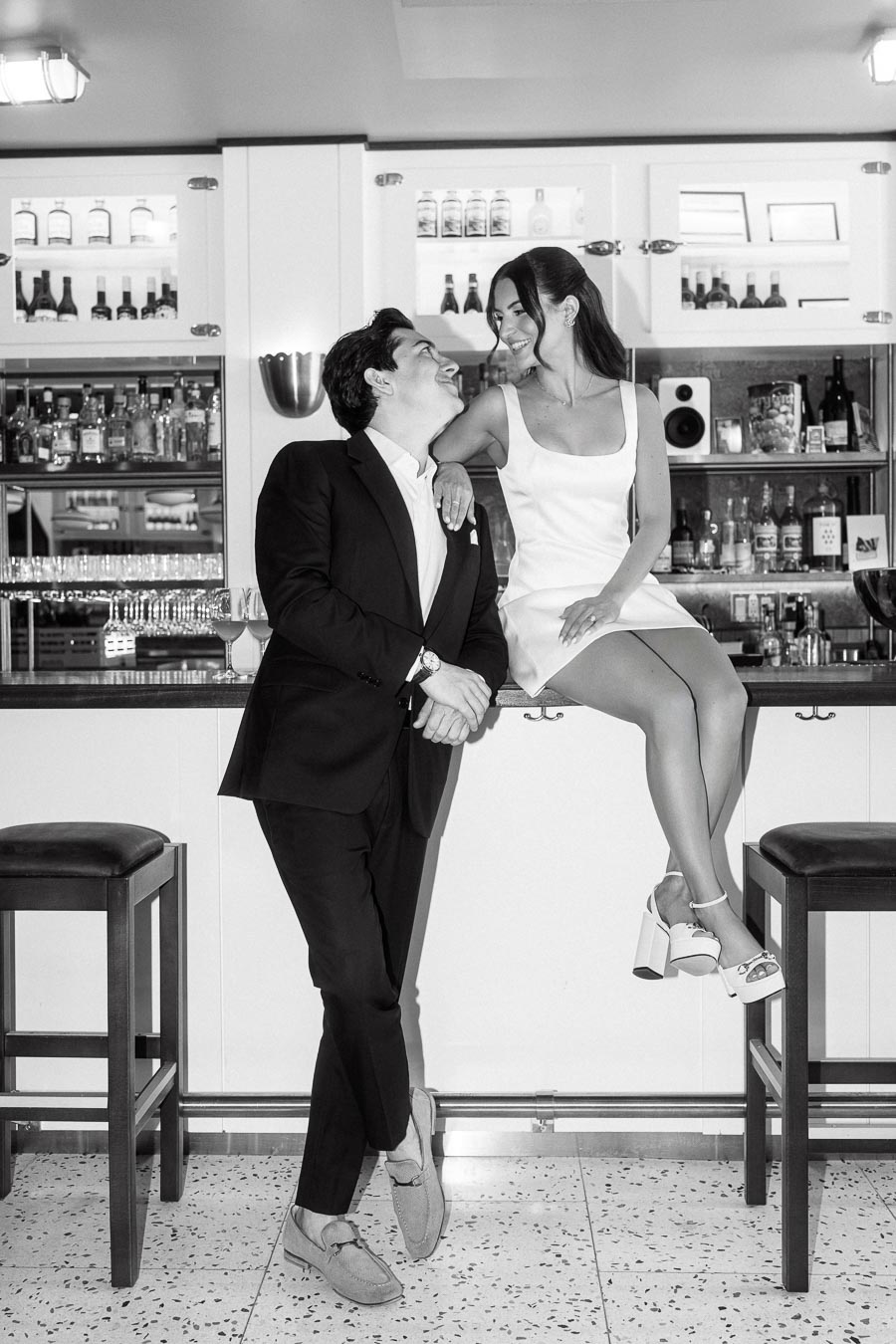 A couple in formal attire smiles and gazes at each other in a modern bar, with the woman sitting on the counter and the man leaning beside her.