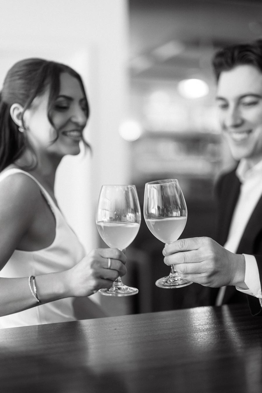 A smiling couple clinks wine glasses together across a bar table, enjoying a romantic and joyful engagement moment in Ontario, Canada.