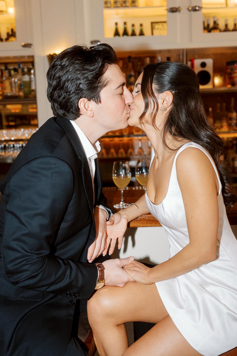 A couple in formal attire shares a kiss while sitting at a bar with two glasses of wine in the background, Ontario, Canada.