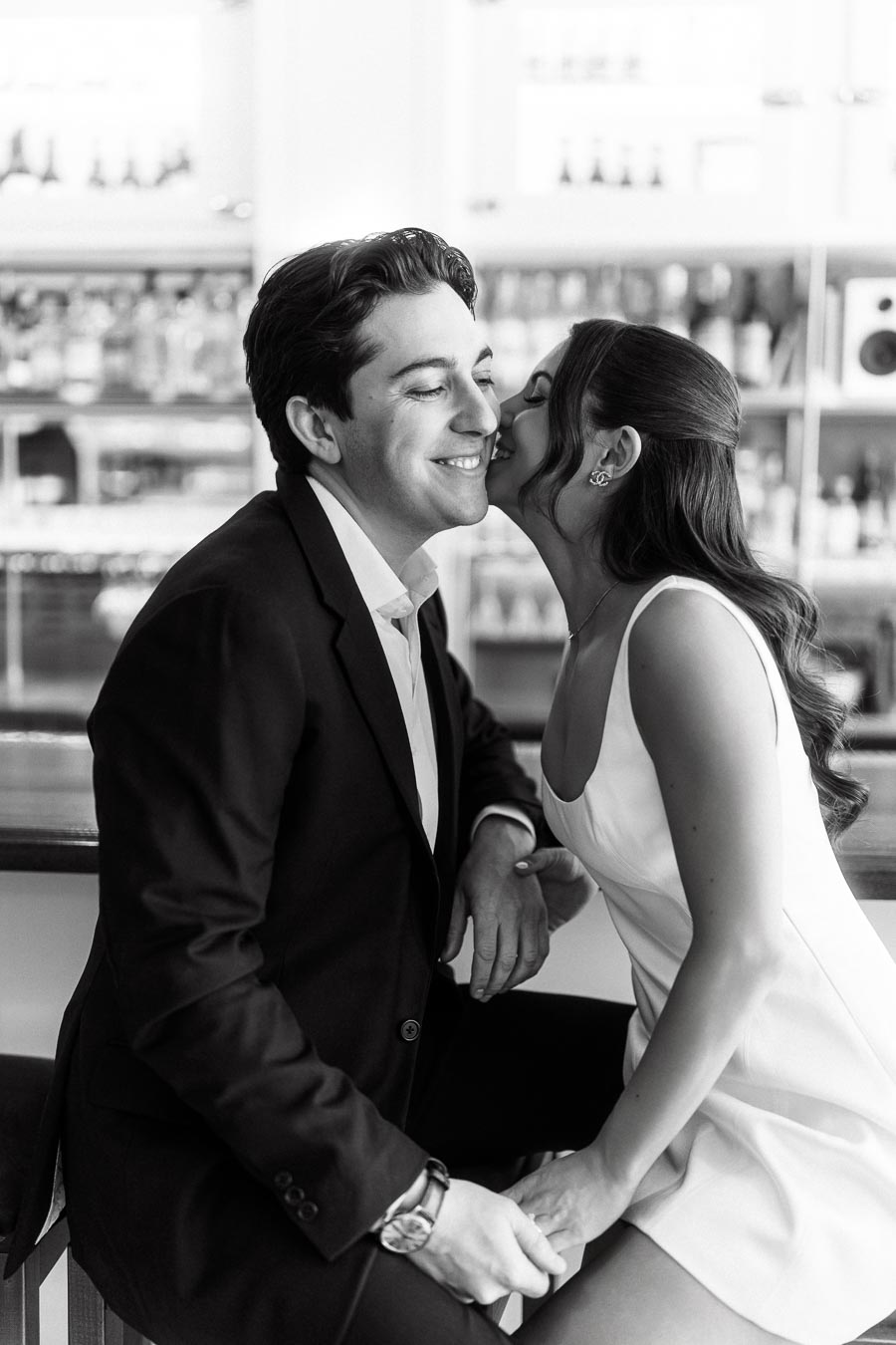 A couple sits closely at a bar, the woman in a white dress kisses the man's cheek, both smiling, with bottles visible in the background.