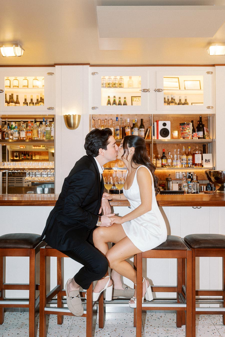 A couple dressed in formal attire shares a kiss while seated on bar stools in a warmly lit indoor bar setting, with bottles and glasses in the background.