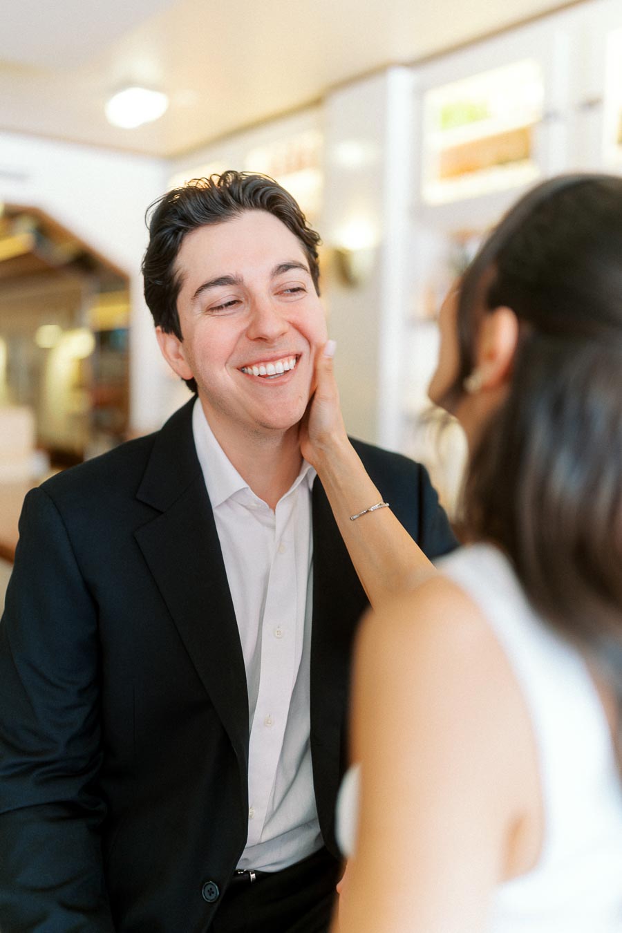 Smiling man in a suit being gently touched on the cheek by a woman in a white dress, both seated in a softly lit indoor setting.