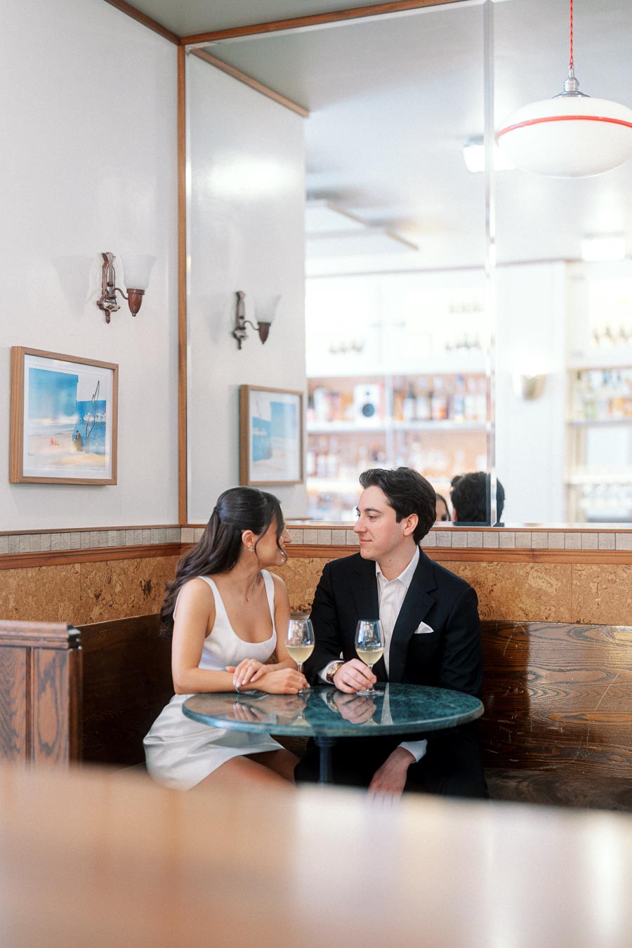 A couple sits closely together at a cozy bar table, smiling and holding wine glasses, with soft indoor lighting and modern decor.
