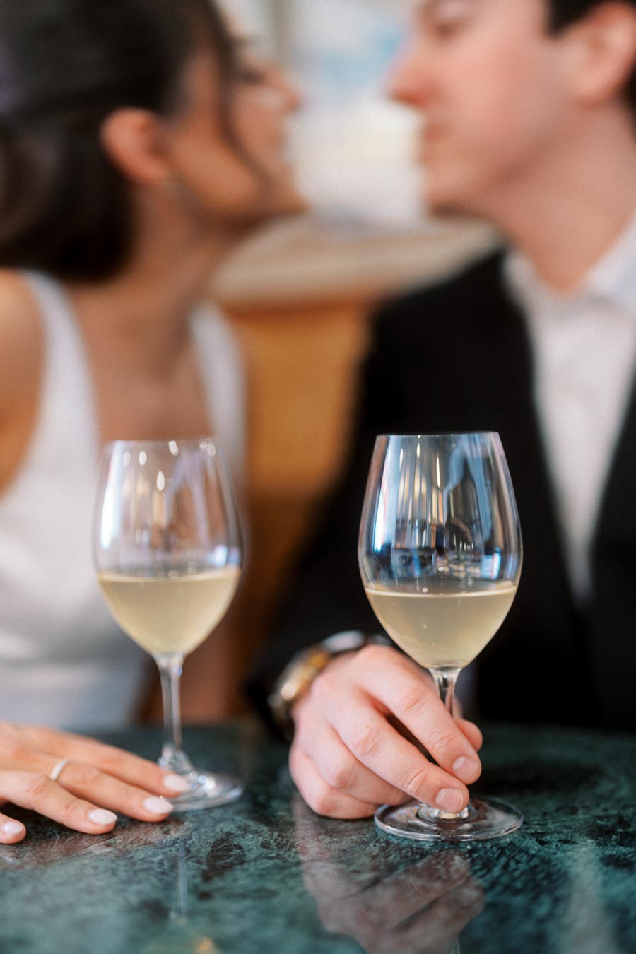 Close-up of two wine glasses on a green marble table, with a couple in the background sharing an intimate moment at a bar in Ontario.