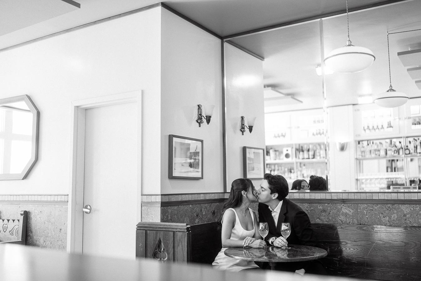 A couple sits at a round table in a stylish indoor bar, sharing a kiss and holding hands, with wine glasses in front of them.