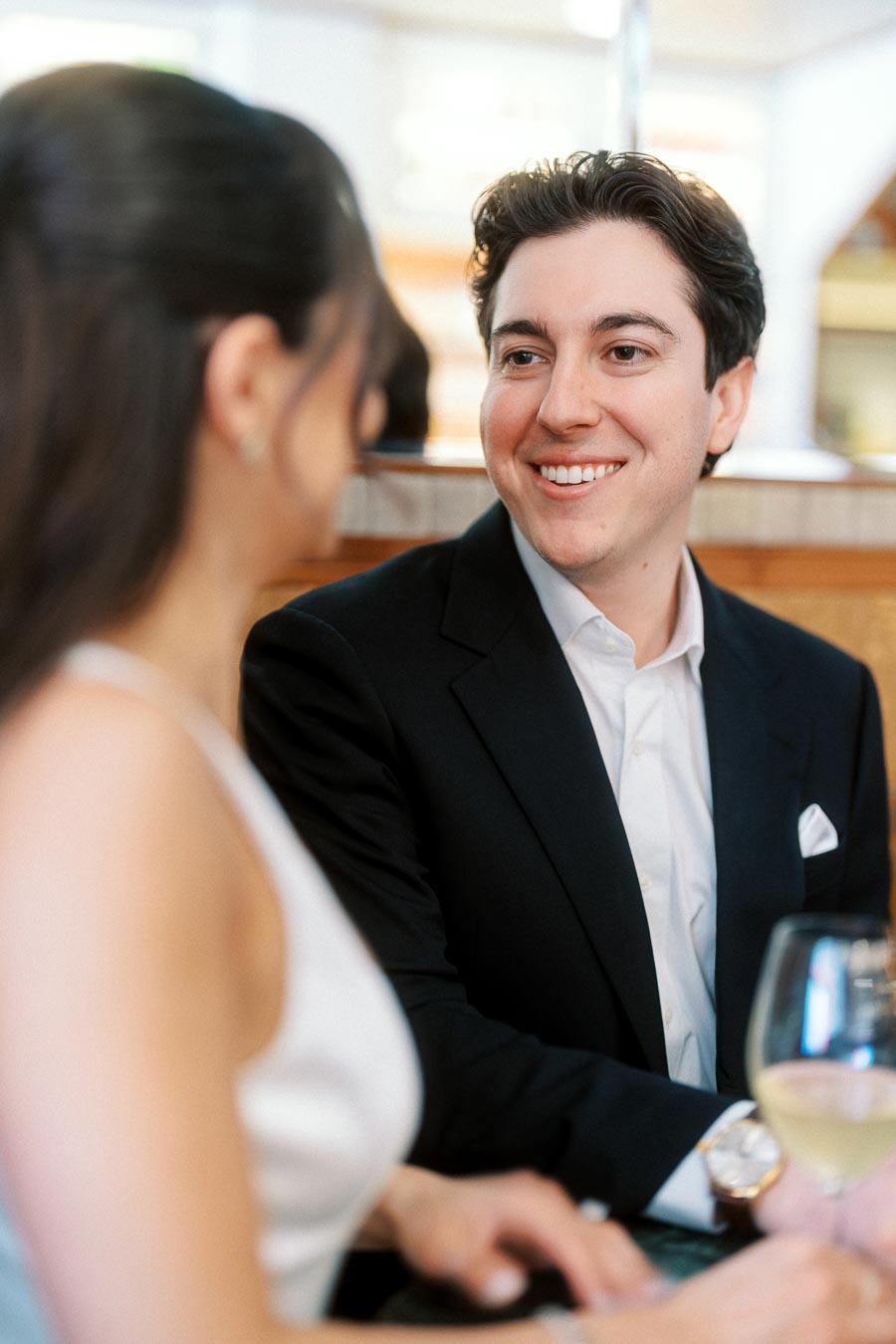 A man in a dark suit smiles at a woman in a white dress as they sit together at a bar table with wine glasses, in Ontario, Canada.