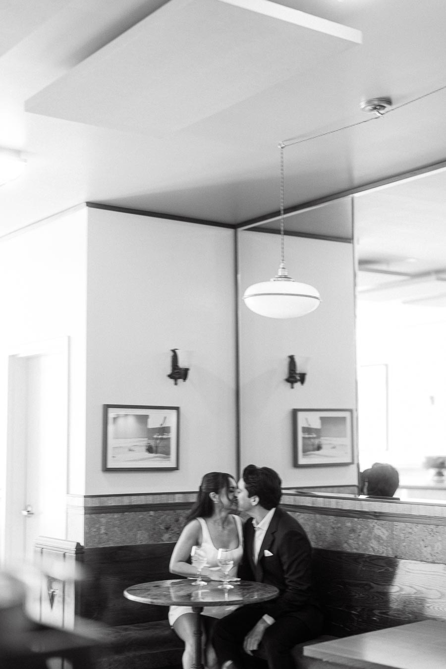 A couple sits closely at a round table in a stylish indoor bar, sharing a kiss with drinks in front of them, captured in black and white.