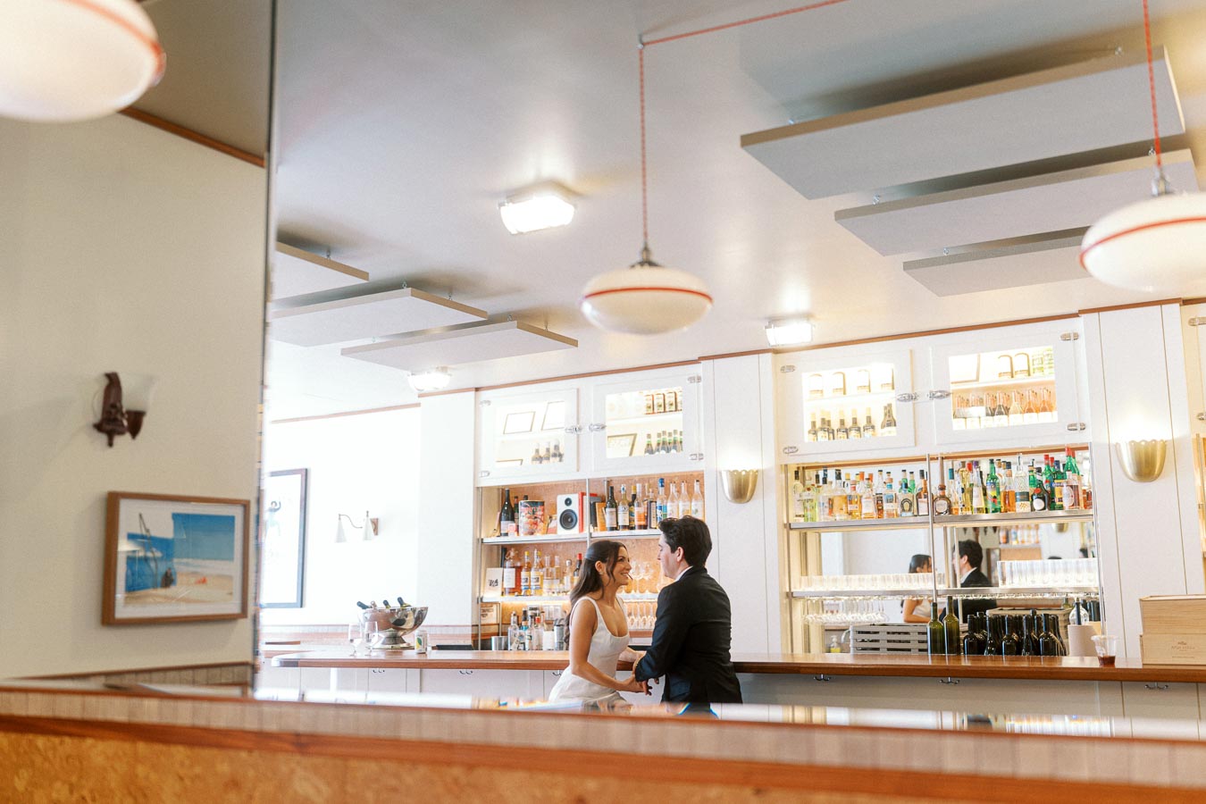A couple dressed in formal attire sits at a modern bar, laughing and holding hands, with shelves of bottles behind them in Ontario, Canada.